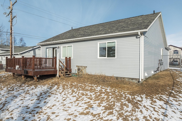 snow covered back of property featuring fence, a deck, and roof with shingles