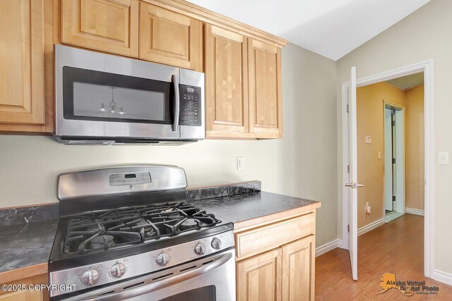 kitchen with dark countertops, light brown cabinetry, vaulted ceiling, light wood-style floors, and stainless steel appliances