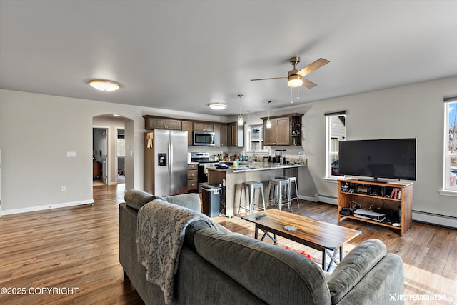 living area with ceiling fan, arched walkways, a baseboard heating unit, baseboards, and light wood-style floors