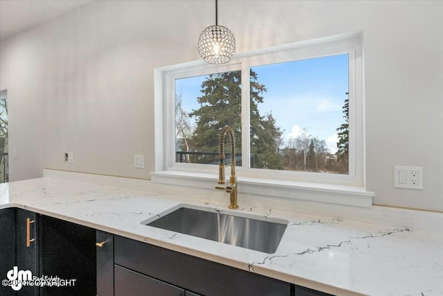 kitchen with dark cabinets, a sink, hanging light fixtures, and light stone counters