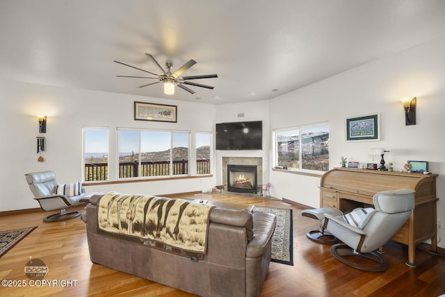 living room with wood finished floors, baseboards, a warm lit fireplace, and ceiling fan