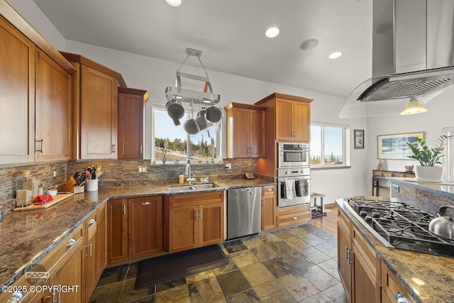 kitchen featuring decorative backsplash, brown cabinets, stainless steel appliances, wall chimney exhaust hood, and a sink