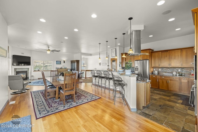 kitchen with a breakfast bar area, brown cabinetry, island exhaust hood, stainless steel fridge with ice dispenser, and tasteful backsplash