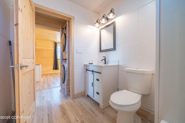 bathroom with wood walls, toilet, stacked washing maching and dryer, wood finished floors, and vanity