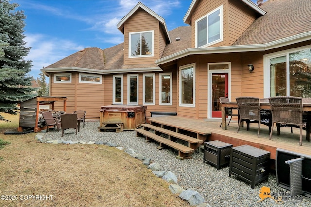 back of property with a deck, outdoor dining space, roof with shingles, and a hot tub