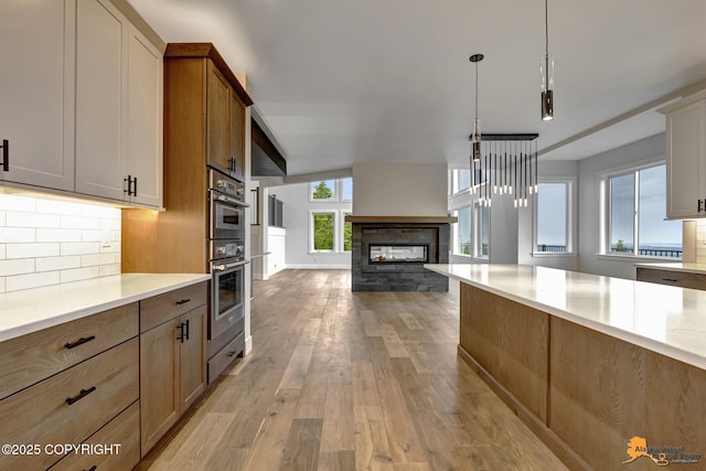 kitchen with backsplash, a multi sided fireplace, double oven, light countertops, and light wood-style flooring