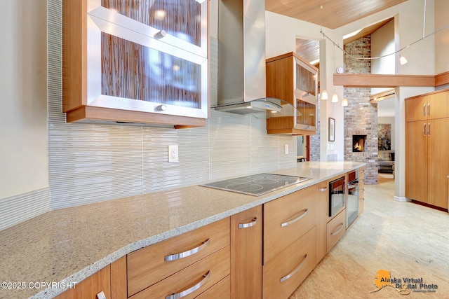 kitchen with light stone countertops, black electric stovetop, glass insert cabinets, wall chimney range hood, and backsplash