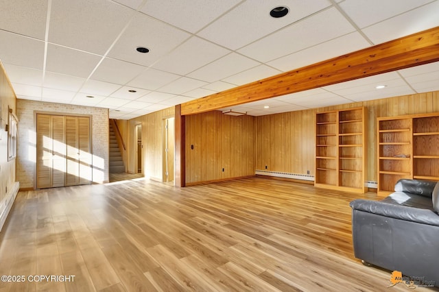 unfurnished living room featuring stairway, a baseboard radiator, a drop ceiling, light wood-style floors, and wood walls