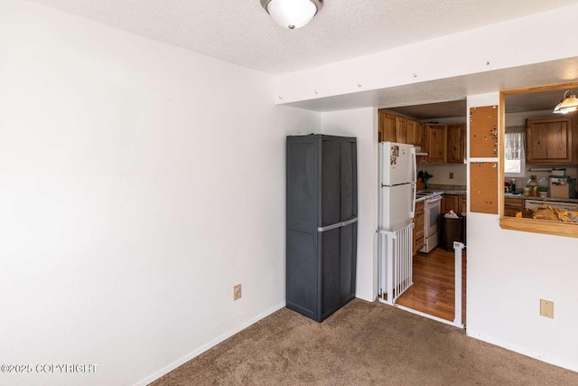 kitchen featuring a textured ceiling, white appliances, brown cabinetry, baseboards, and light colored carpet