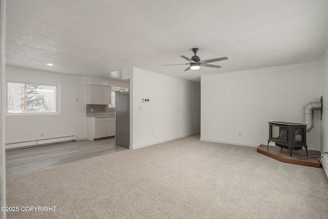 unfurnished living room with baseboards, a baseboard radiator, a wood stove, ceiling fan, and light colored carpet