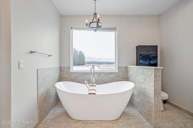 full bath with a freestanding bath, a notable chandelier, wainscoting, tile walls, and toilet