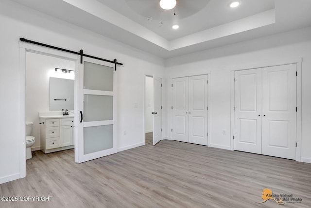 unfurnished bedroom featuring two closets, ensuite bath, a barn door, light wood-style floors, and a raised ceiling