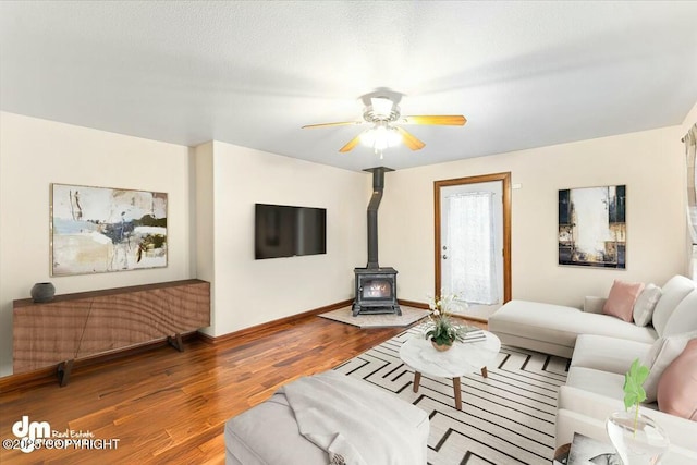 living room with ceiling fan, baseboards, a wood stove, and wood finished floors