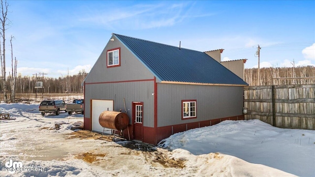view of snowy exterior with an outdoor structure, fence, metal roof, a detached garage, and heating fuel