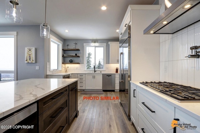 16074 Mountain Air Dr, Anchorage, AK 99516 - kitchen featuring wall chimney exhaust hood, stainless steel appliances, backsplash, light wood-style floors, and hanging light fixtures