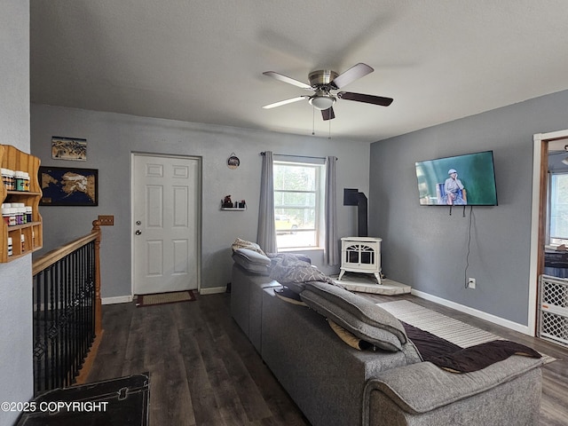 living area with a wood stove, a ceiling fan, dark wood finished floors, and baseboards