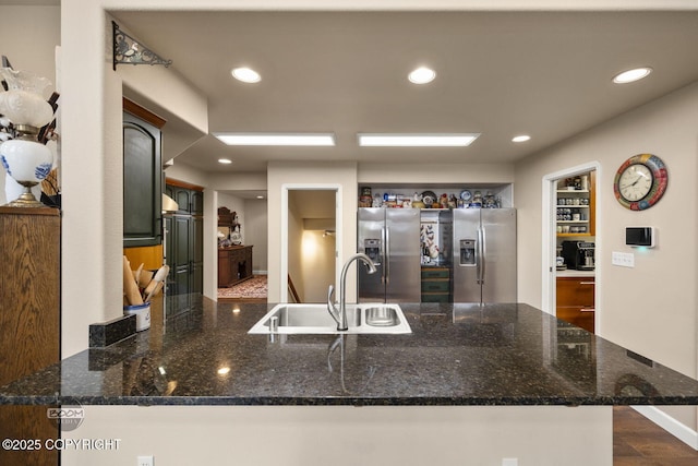 kitchen featuring stainless steel refrigerator with ice dispenser, a sink, a peninsula, recessed lighting, and dark stone counters