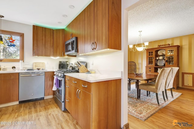kitchen with brown cabinetry, light countertops, light wood-style flooring, appliances with stainless steel finishes, and a notable chandelier