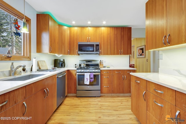 kitchen with brown cabinetry, appliances with stainless steel finishes, hanging light fixtures, a sink, and light wood-style flooring
