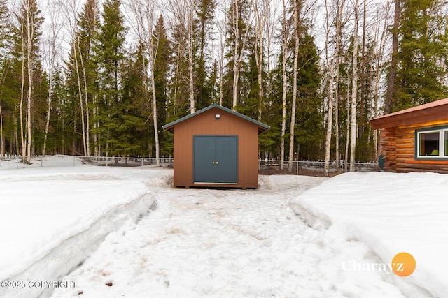 snow covered structure with a storage shed, an outbuilding, fence, and a wooded view