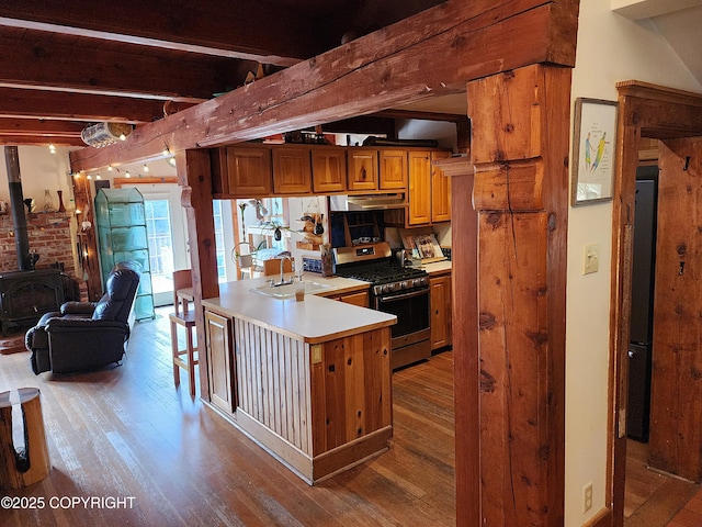 kitchen with brown cabinets, under cabinet range hood, beamed ceiling, light wood-type flooring, and stainless steel gas range