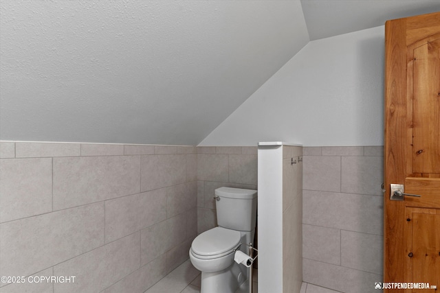 3915 Hiland Rd, Eagle River, AK 99577 - bathroom featuring tile walls, vaulted ceiling, toilet, wainscoting, and tile patterned flooring
