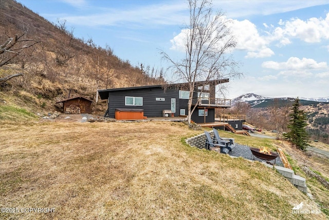 rear view of house featuring a fire pit, a deck with mountain view, a lawn, a hot tub, and an outbuilding