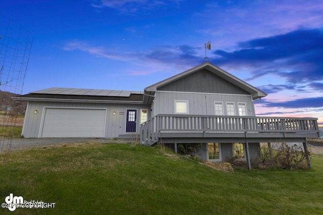 back of house at dusk with an attached garage, a wooden deck, dirt driveway, solar panels, and a yard