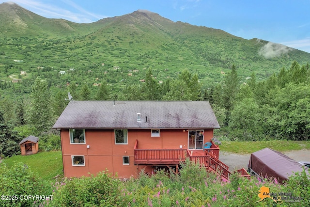 rear view of house with a deck with mountain view, an outbuilding, a wooded view, and a shingled roof