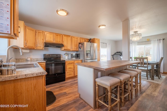 kitchen with appliances with stainless steel finishes, a sink, under cabinet range hood, a breakfast bar, and dark wood-style flooring