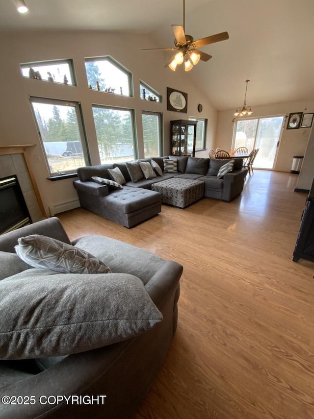 living room featuring a tiled fireplace, wood finished floors, high vaulted ceiling, a baseboard radiator, and ceiling fan with notable chandelier