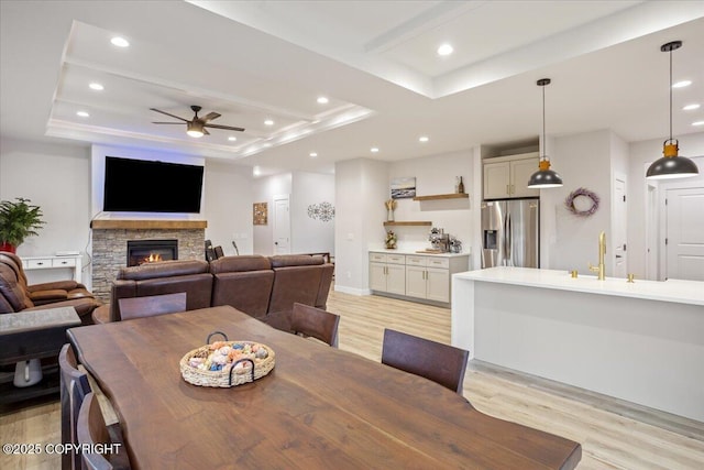 dining room with ceiling fan, a raised ceiling, a stone fireplace, recessed lighting, and light wood-type flooring