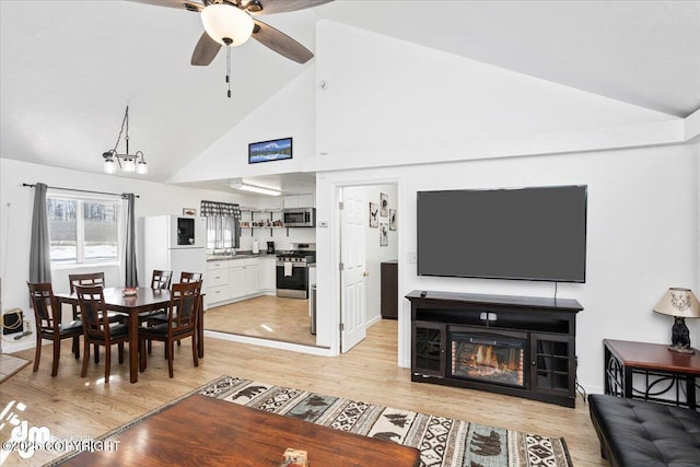living room with high vaulted ceiling, a glass covered fireplace, ceiling fan with notable chandelier, and light wood-style floors