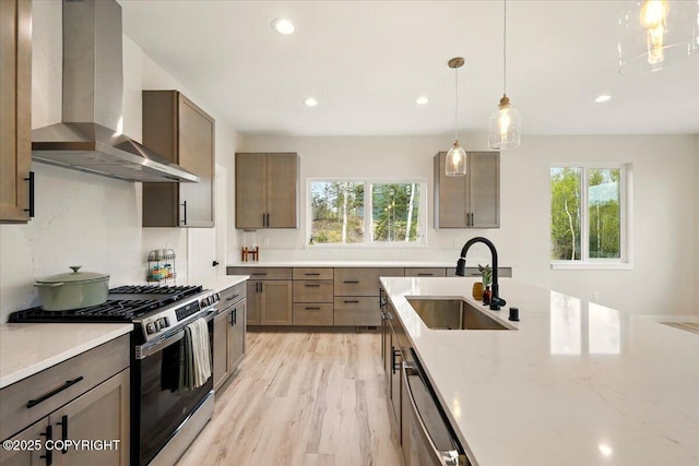 kitchen with wall chimney range hood, appliances with stainless steel finishes, light stone counters, a sink, and light wood-style flooring