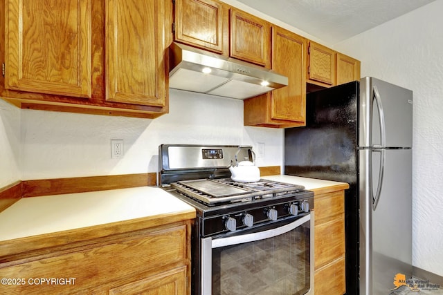 kitchen with under cabinet range hood, appliances with stainless steel finishes, light countertops, brown cabinetry, and a textured wall