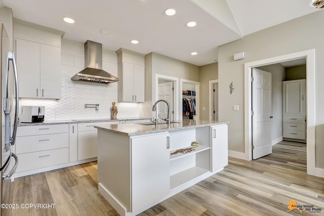 kitchen featuring wall chimney exhaust hood, a sink, light wood finished floors, decorative backsplash, and a center island with sink
