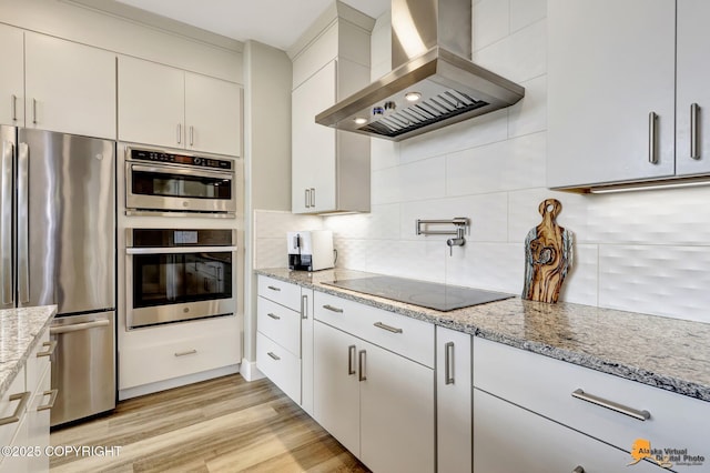 kitchen with stainless steel appliances, wall chimney exhaust hood, tasteful backsplash, light wood-style floors, and light stone countertops