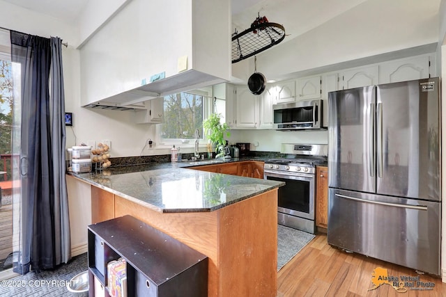 kitchen with appliances with stainless steel finishes, a sink, a peninsula, dark stone countertops, and light wood-type flooring
