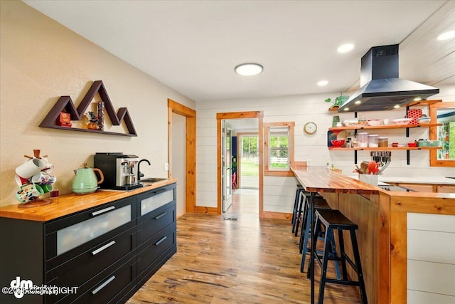 kitchen featuring wooden counters, island exhaust hood, dark cabinets, a sink, and light wood-type flooring