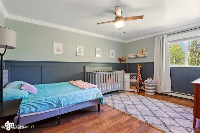 bedroom with ornamental molding, wood finished floors, wainscoting, a baseboard radiator, and ceiling fan