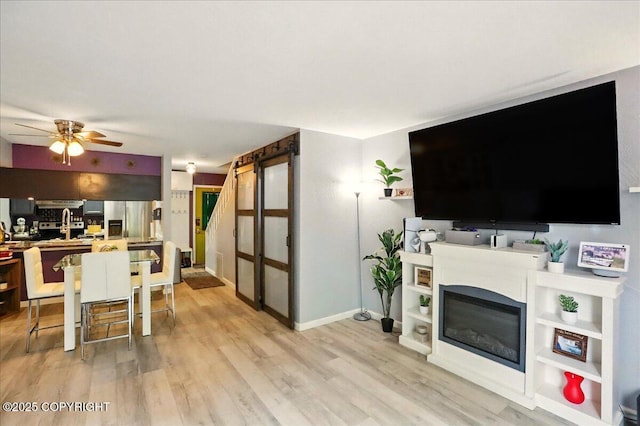 living area featuring ceiling fan, light wood-style floors, a glass covered fireplace, and baseboards