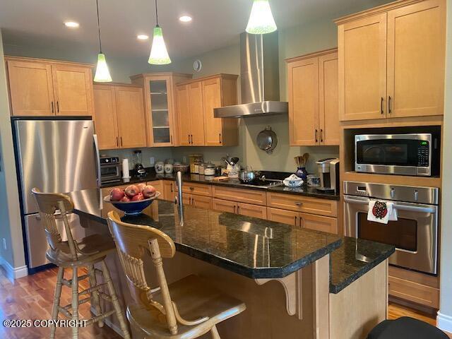 2295 N Filly Cir, Wasilla, AK 99654 - kitchen featuring stainless steel appliances, wall chimney exhaust hood, wood finished floors, glass insert cabinets, and a center island with sink