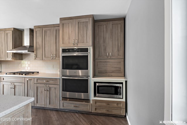 8410 E 20th Ave, Anchorage, AK 99504 - kitchen featuring appliances with stainless steel finishes, wall chimney range hood, decorative backsplash, dark wood-style flooring, and light stone countertops