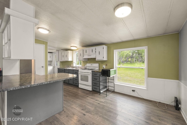 1801 W That Rd, Wasilla, AK 99623 - kitchen with white range with gas cooktop, a wainscoted wall, dark wood-style floors, under cabinet range hood, and white cabinetry