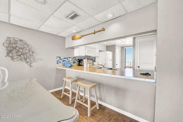 kitchen with a drop ceiling, a breakfast bar, white appliances, white cabinets, and tile floors