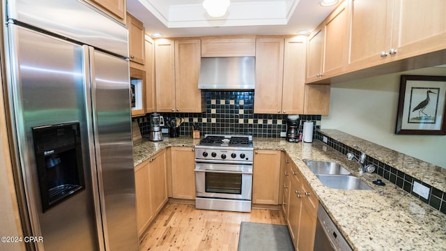 kitchen featuring wall chimney range hood, a sink, light brown cabinetry, light wood finished floors, and appliances with stainless steel finishes