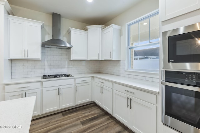 kitchen with dark wood-type flooring, white cabinets, wall chimney exhaust hood, decorative backsplash, and appliances with stainless steel finishes