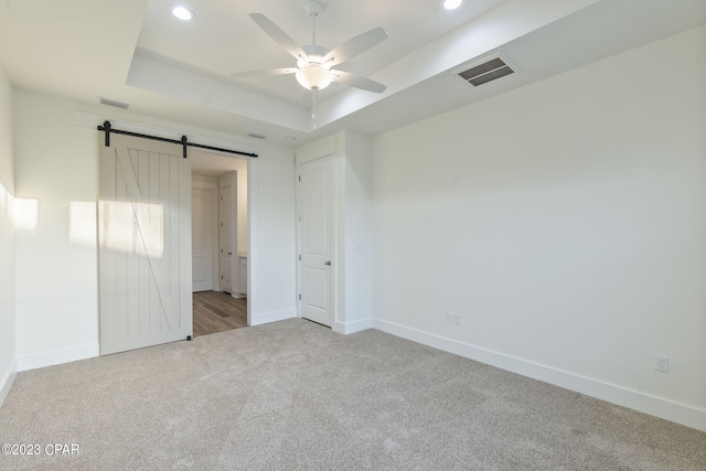 unfurnished bedroom with ceiling fan, a barn door, light colored carpet, and a tray ceiling