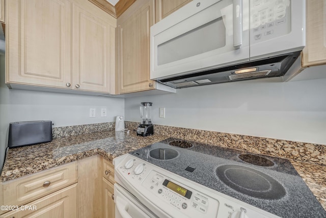 kitchen featuring light brown cabinetry, dark stone counters, and white appliances