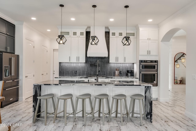 kitchen featuring white cabinetry, backsplash, appliances with stainless steel finishes, custom range hood, and an island with sink